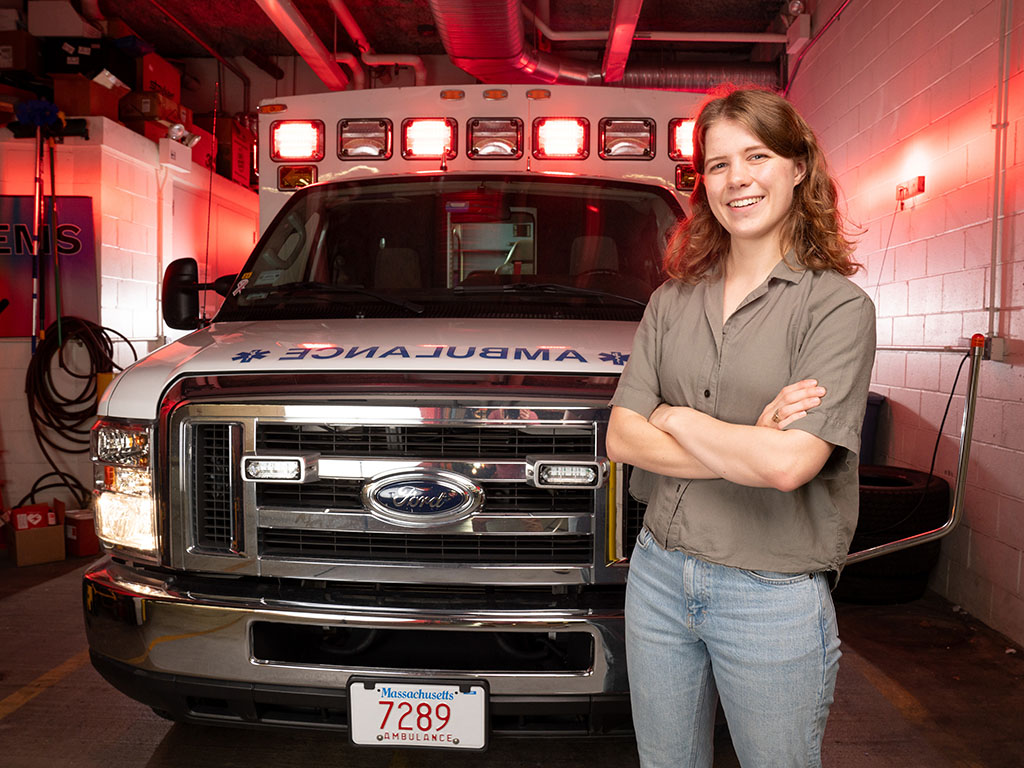 Abigail Schipper smiles with arms crossed while standing in front of a parked MIT ambulance with red lights on.
