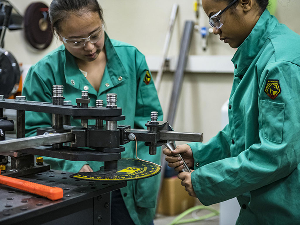 Two young women in teal safety jumpsuits work together in a machine shop