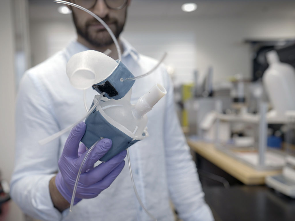 A person in a lab coat holds a blue and white 3d-printed heart replica, a soft and flexible shell with fabricated sleeves, in their gloved hand.