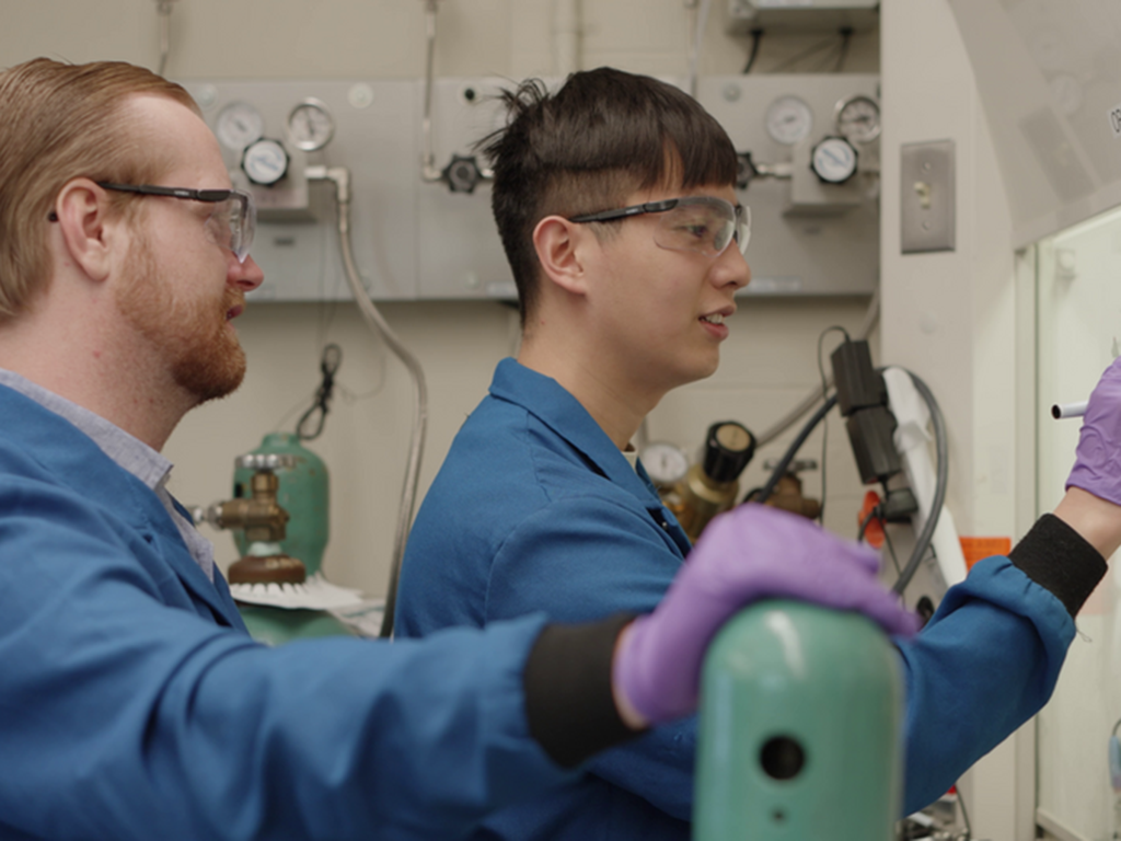 Two researchers dressed in lab attire review writing on a dry erase board
