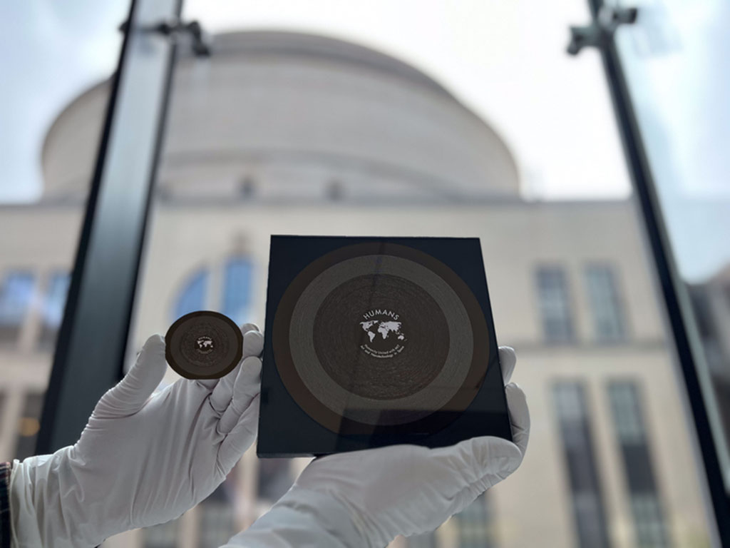 Two brown-gold nanowafers held up by white-gloved hands in front of a window with MIT's Great Dome in the background. The lunar HUMANS wafer is on the left, it is 2 inches in diameter and features an equal-area projection world map in the center. The HUMA