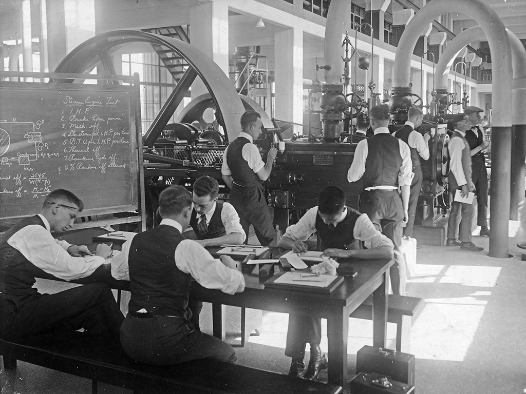 A black and white image of men working at tables and on a steam engine