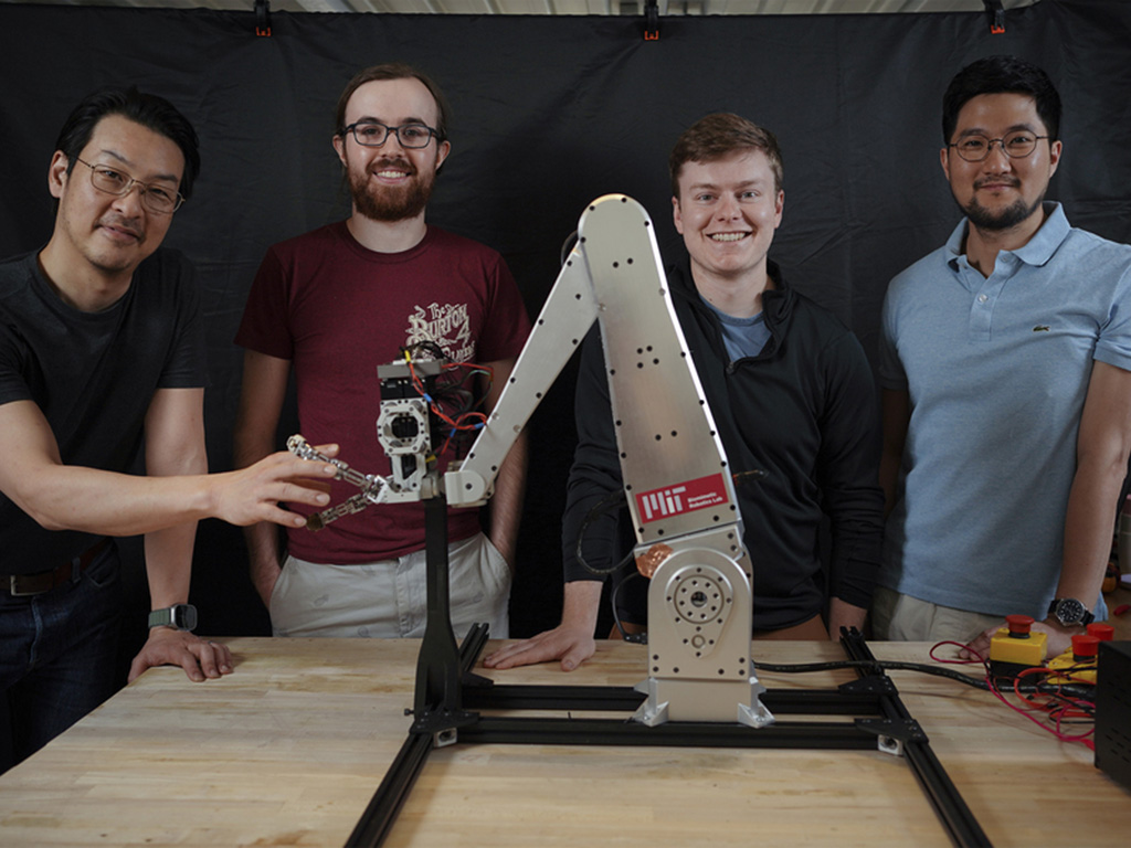 Sangbae Kim, Elijah Stanger-Jones, Andrew SaLoutos, and Hongmin Kim pose for a portrait as they stand behind a metal robot gripper that resembles an excavator. Sangbae Kim’s fingers lace with the gripper.