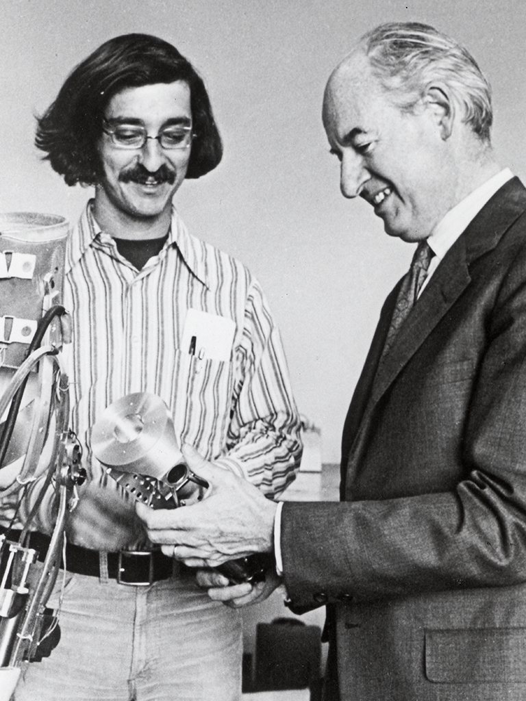 A black and white image of Robert Mann and Woodie Flowers, smiling at something in Flowers' hands.