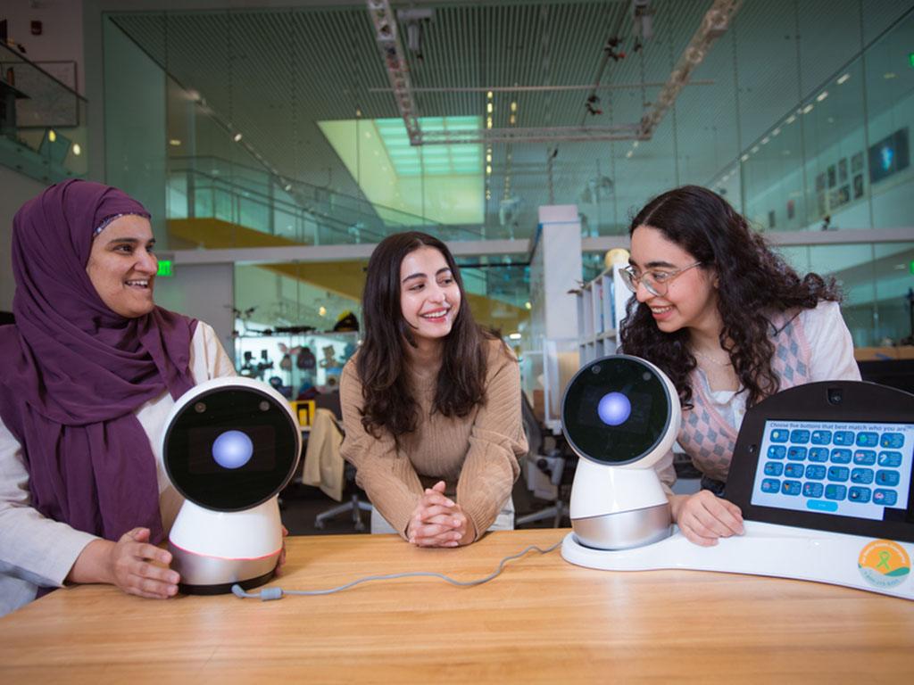 Sharifa Alghowinem with head scarf and Deim Alfozan and Tasneem Burghleh, both with long dark brown hair, lean on a table bearing two Jibo personal robots, which look like spheres with large, single eyes."