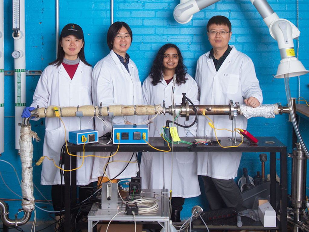 Chuwei Zhang, Sili Deng, Maanasa Bhat, and Jianan Zhang stand in white lab coats behind a metal cylinder wrapped in insulation and festooned with wires
